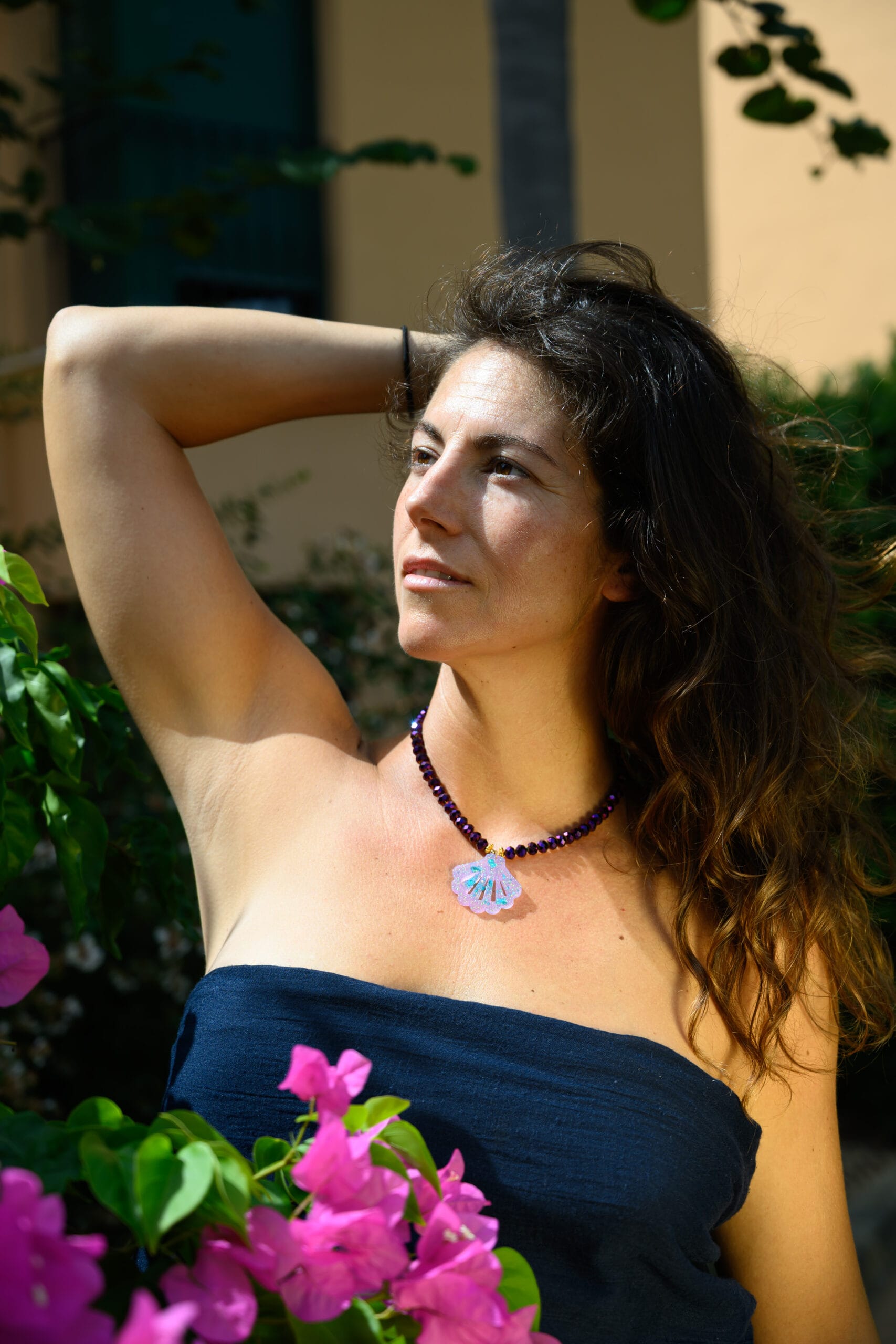Collar de cuentas moradas facetadas con colgante de concha puesto en el cuello, modelo luce con pelo recogido y brazo levantado