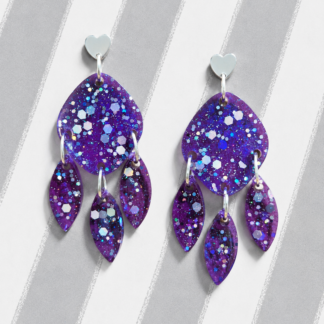 Pendientes morados Lavanda Party de resina con corazón plateado, purpurina holográfica y tres pétalos colgantes sobre fondo de rayas grises